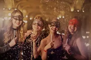 Fire kvinder i masker og festkjoler fejrer til en temafest med champagne og konfetti i en smukt oplyst balsal.